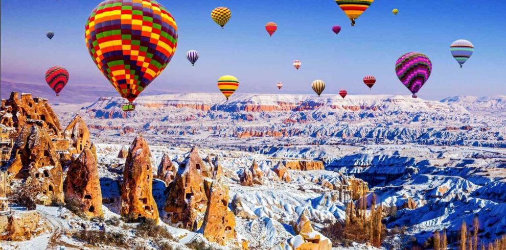 Vibrant hot air balloons soar above the snow-dusted rock formations of Cappadocia, Turkey, on a clear winter day.