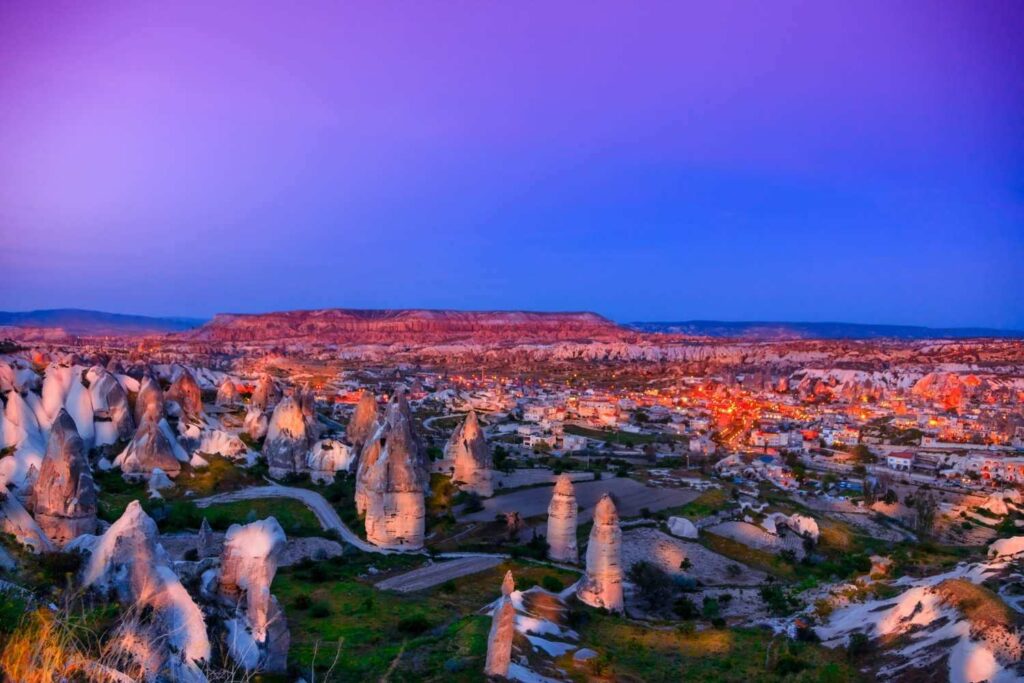 Cappadocia's rocky terrain illuminated by town lights at twilight, with a purple sky adding a magical ambiance.