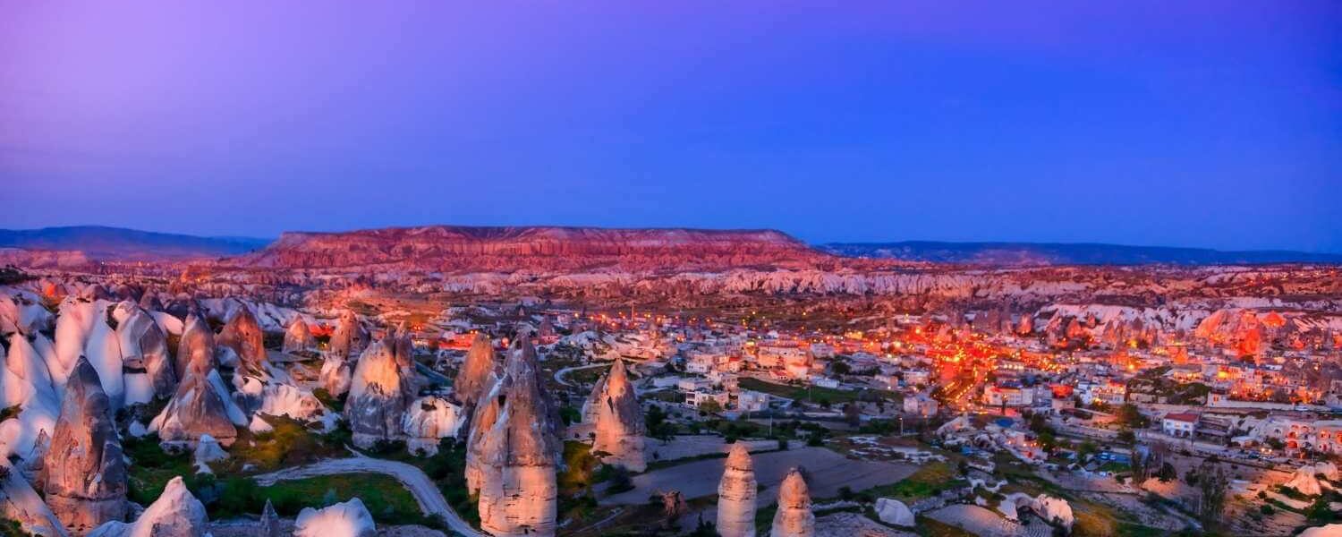 Cappadocia's rocky terrain illuminated by town lights at twilight, with a purple sky adding a magical ambiance.