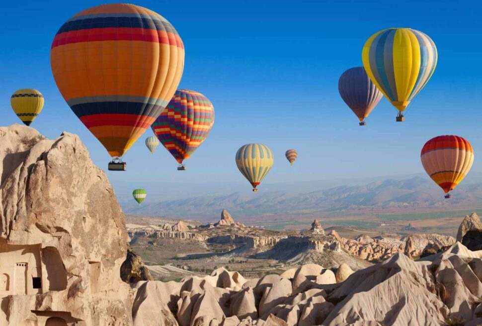 Vibrant hot air balloons floating over the rocky landscape of Cappadocia, Turkey, under a clear blue sky.