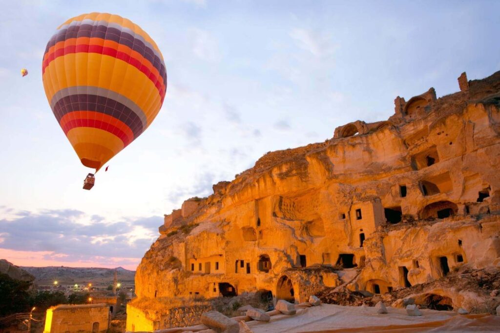 Striped hot air balloon floats near ancient cave houses in Cappadocia, Turkey, during sunset with a soft sky.