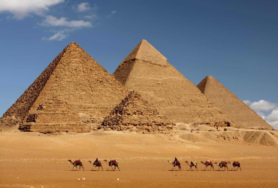 The Pyramids of Giza under a clear sky, with a camel caravan traversing the desert in the foreground.