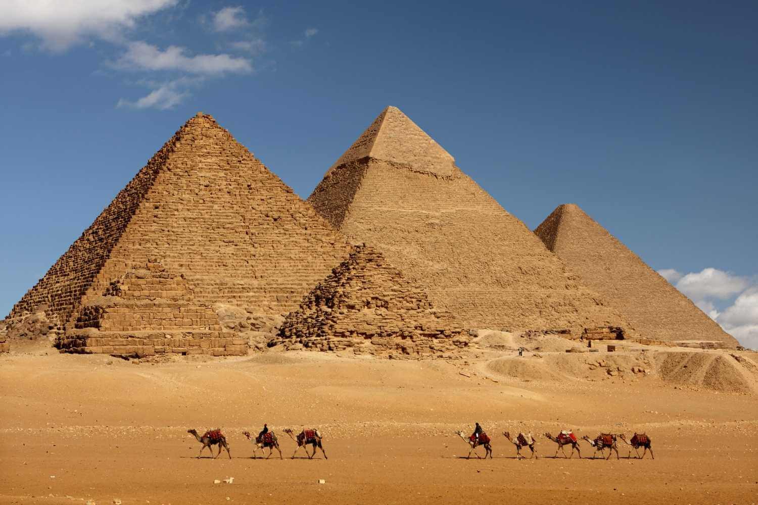 The Pyramids of Giza under a clear sky, with a camel caravan traversing the desert in the foreground.