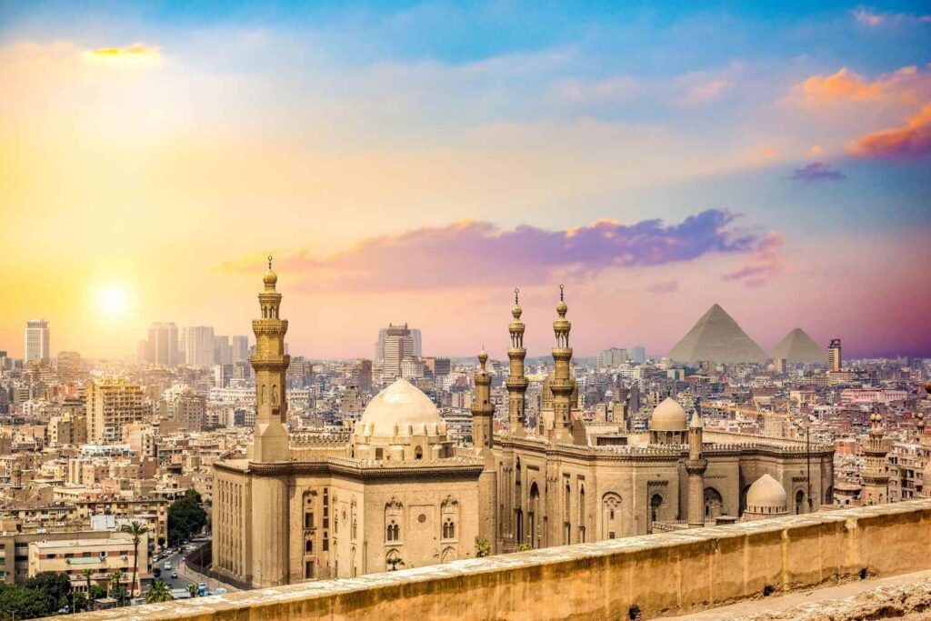 The skyline of Cairo at sunset, featuring historic mosques in the foreground and the Pyramids of Giza in the background.