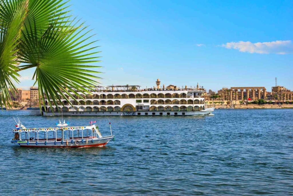 A large cruise ship and a small boat sailing on the Nile River with the Luxor Temple and cityscape in the background.