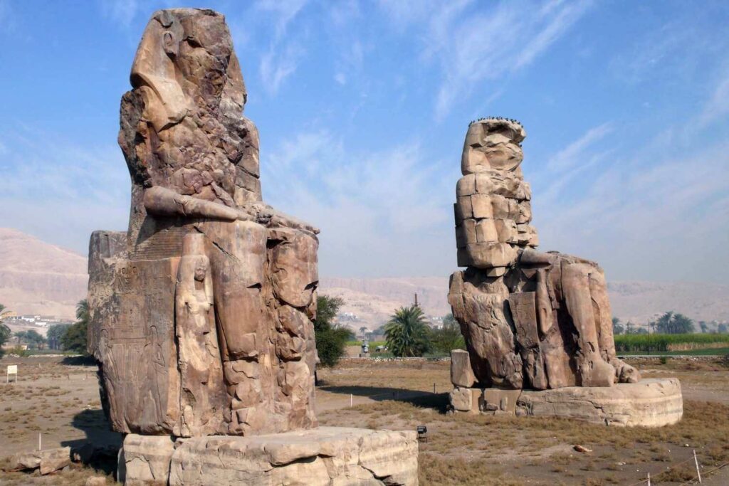 Two massive, weathered stone statues of pharaohs known as the Colossi of Memnon, set in an open landscape under a cloudy sky.