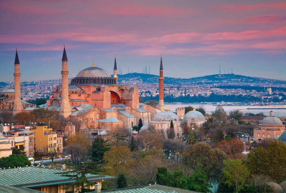 Hagia Sophia in Istanbul at sunset, featuring its domes and minarets with a cityscape and water in the background.