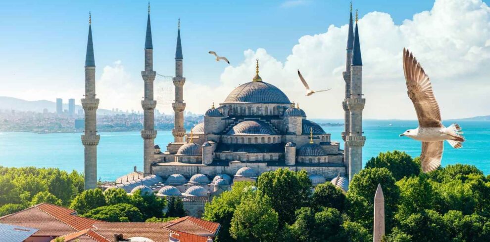 The Blue Mosque in Istanbul, with its domes and six minarets, set against a bright sky and overlooking the sea.