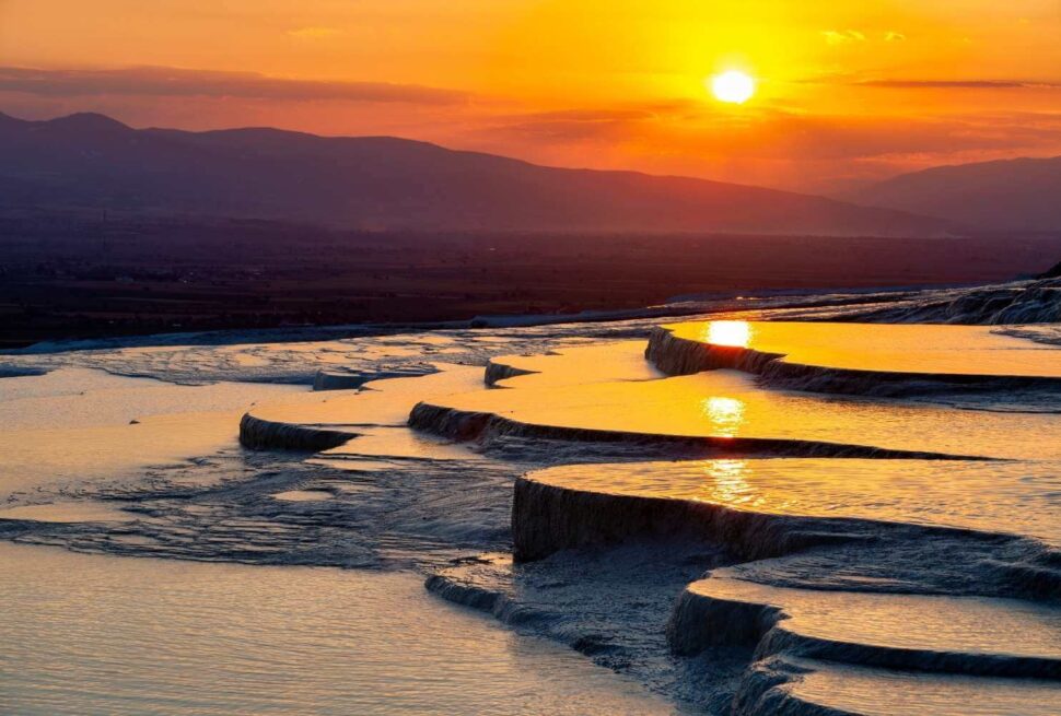 Pamukkale's natural travertine terraces reflecting the vibrant colors of sunset, with mountains in the background.