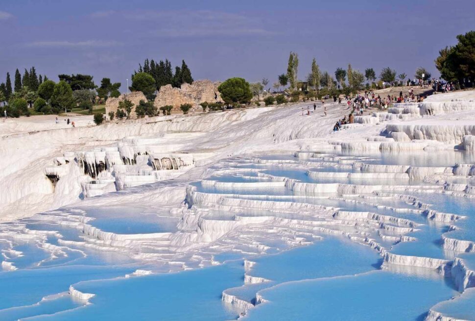 Tourists walking along the white travertine terraces filled with blue thermal water at Pamukkale, Turkey, on a sunny day.