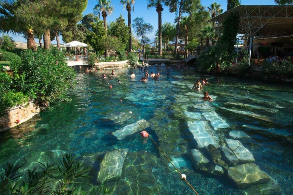 Tourists swimming in the thermal waters of Pamukkale's ancient pool, surrounded by lush greenery and submerged ruins.