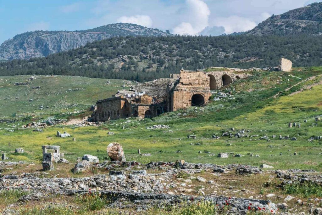 The ancient ruins of Hierapolis, Turkey, set against a lush green landscape and mountainous backdrop under a partly cloudy sky.