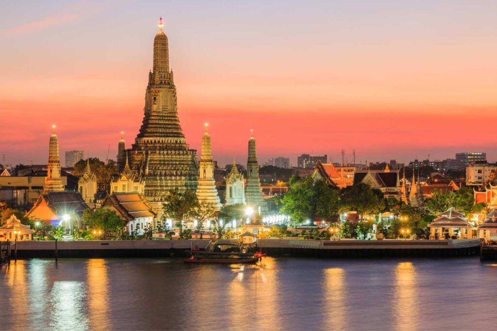 Wat Arun temple in Bangkok illuminated at sunset, reflecting on the Chao Phraya River, with a vibrant sky.