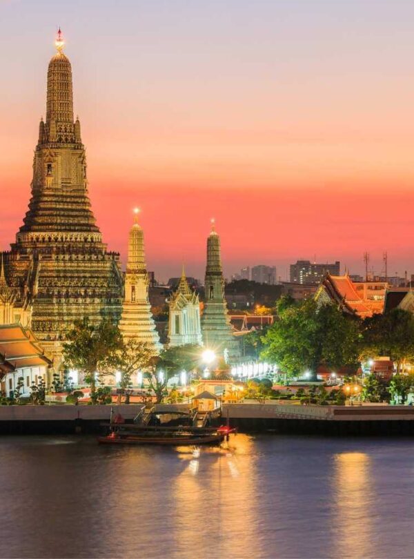 Wat Arun temple in Bangkok illuminated at sunset, reflecting on the Chao Phraya River, with a vibrant sky.