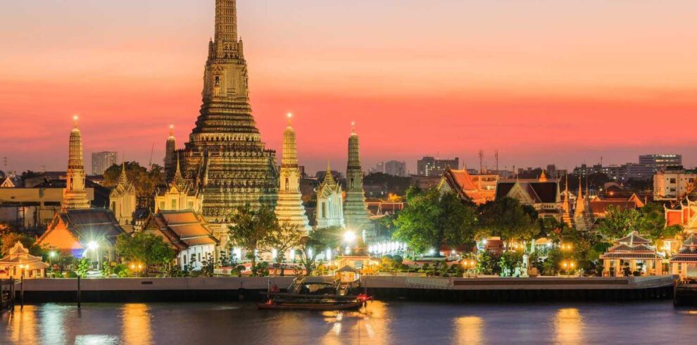 Wat Arun temple in Bangkok illuminated at sunset, reflecting on the Chao Phraya River, with a vibrant sky.