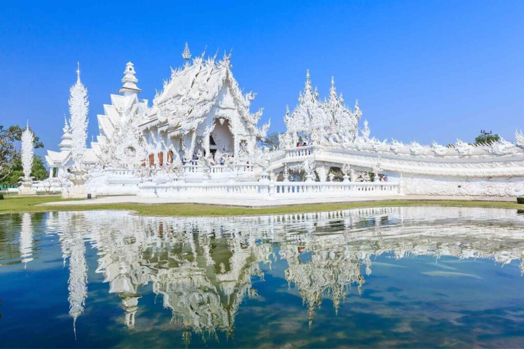 Ornate white temple with intricate carvings, reflecting in a calm pond under a clear blue sky.