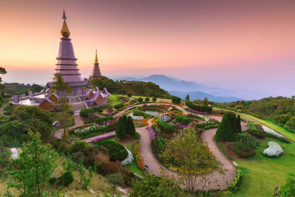 Two towering pagodas surrounded by vibrant gardens on a mountain, set against a colorful sunset sky.