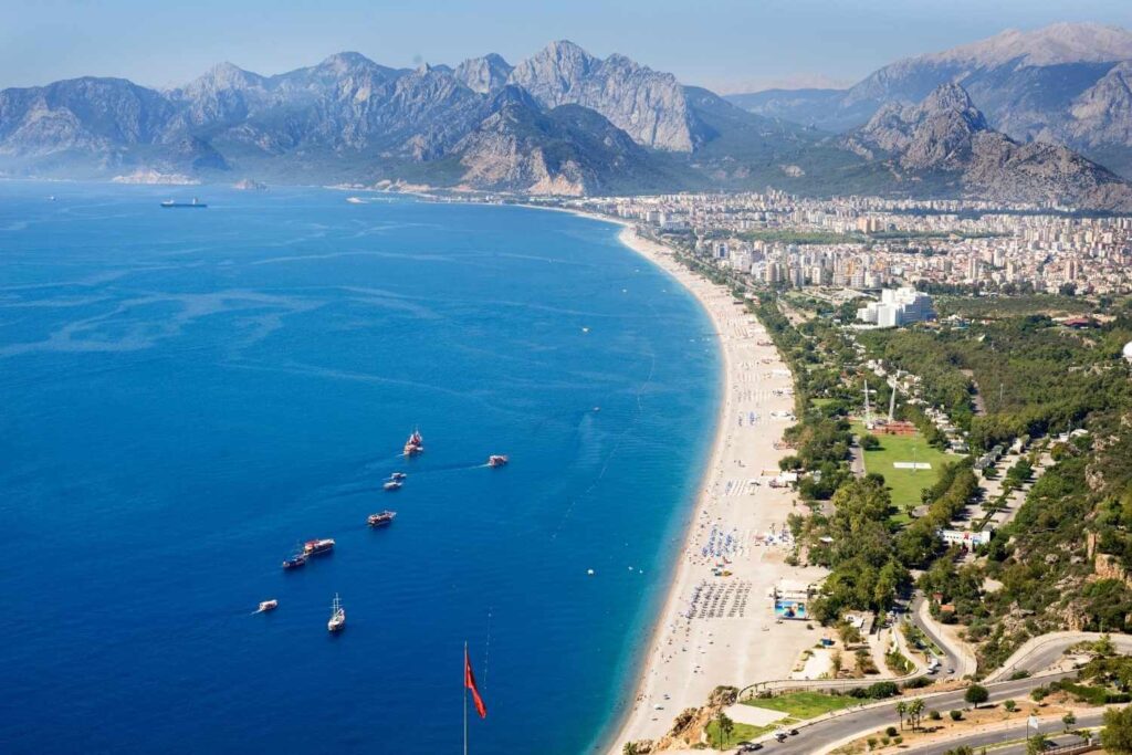 Aerial view of Antalya's Konyaaltı Beach, Turkey, with clear blue water, sandy shoreline, and majestic mountains in the background.