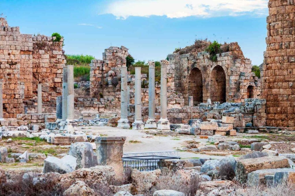 Ancient ruins of Perge in Turkey, featuring stone columns, arched structures, and weathered stone blocks against a blue sky.