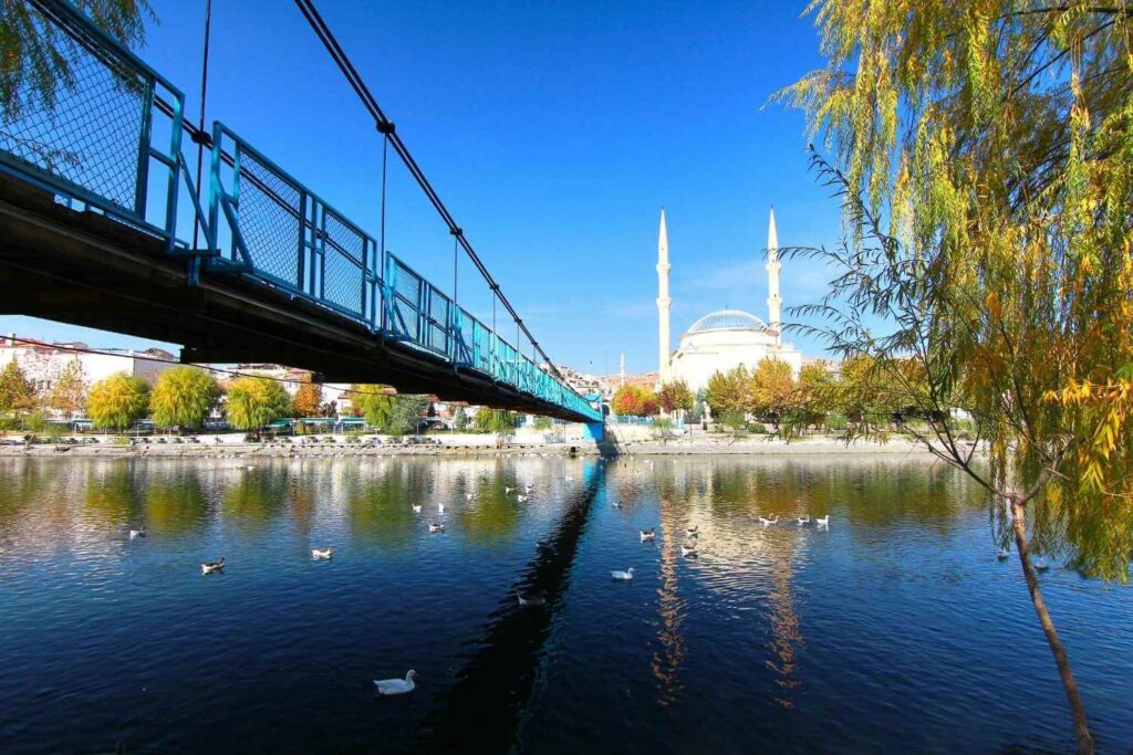 Blue pedestrian bridge over a calm river in Eskişehir, with a mosque and its twin minarets in the background.