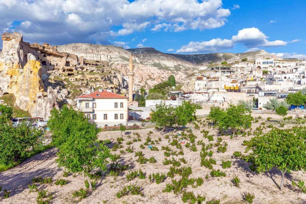 Cavusin Village in Cappadocia with stone houses, cave dwellings, a mosque, and greenery against a mountainous backdrop.