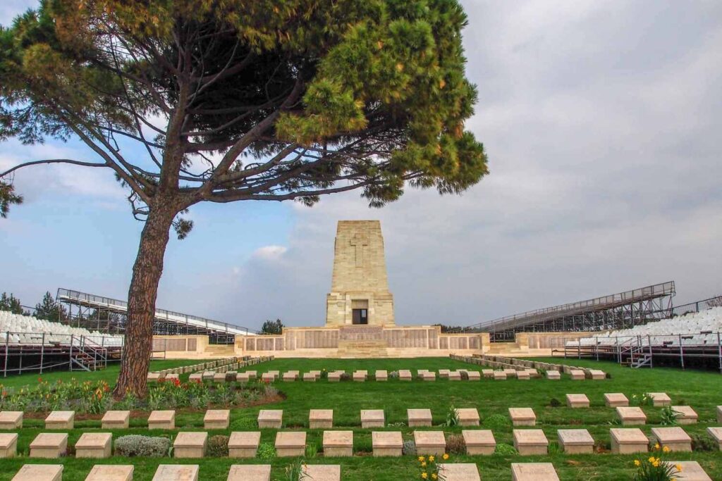 Lone Pine Cemetery with rows of headstones, a tall memorial, and a large tree under a cloudy sky.