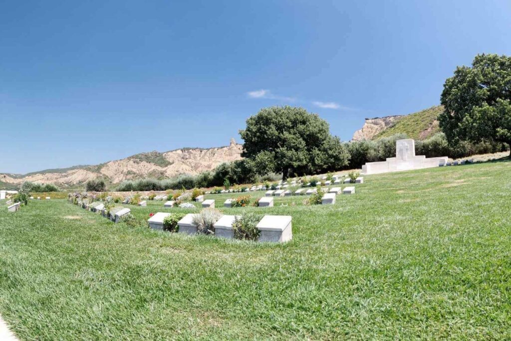 Military cemetery with white headstones on a green lawn, surrounded by hills, trees, and a memorial under a clear sky.