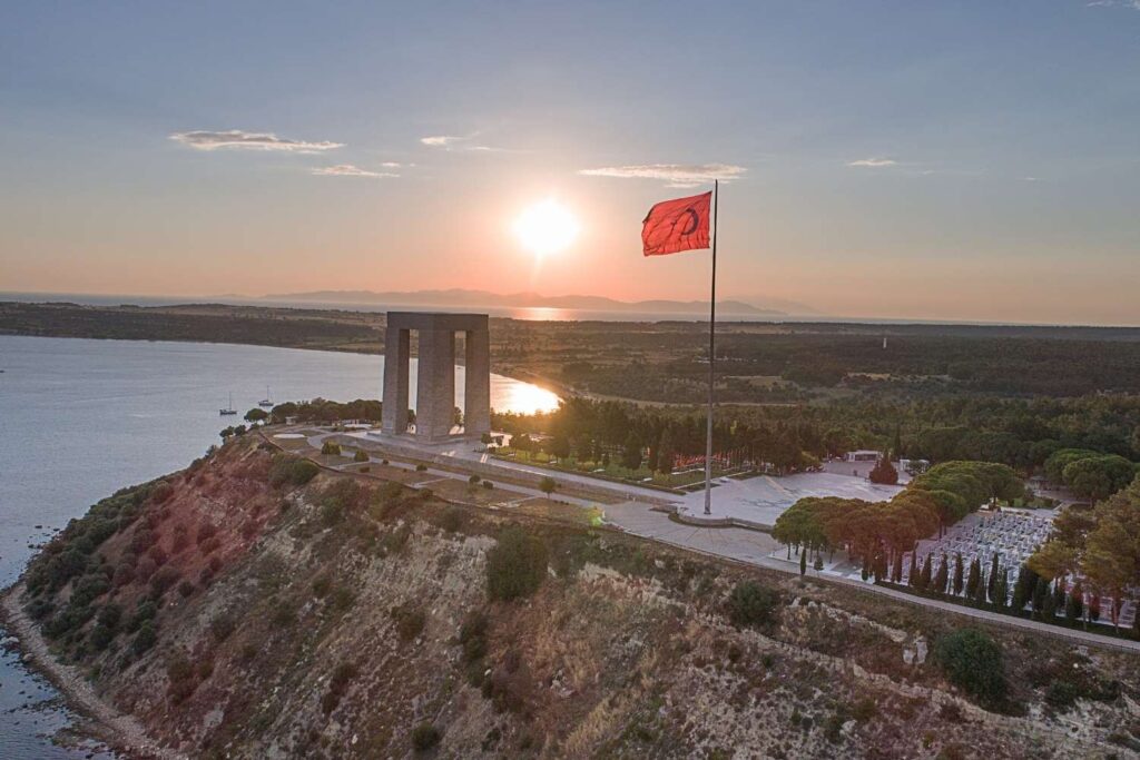 Aerial view of the Çanakkale Martyrs' Memorial at sunset, with a large Turkish flag and coastline in the background.