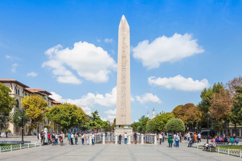 Tourists gather around the ancient Egyptian Obelisk of Theodosius at Istanbul's Sultanahmet Square on a clear day.