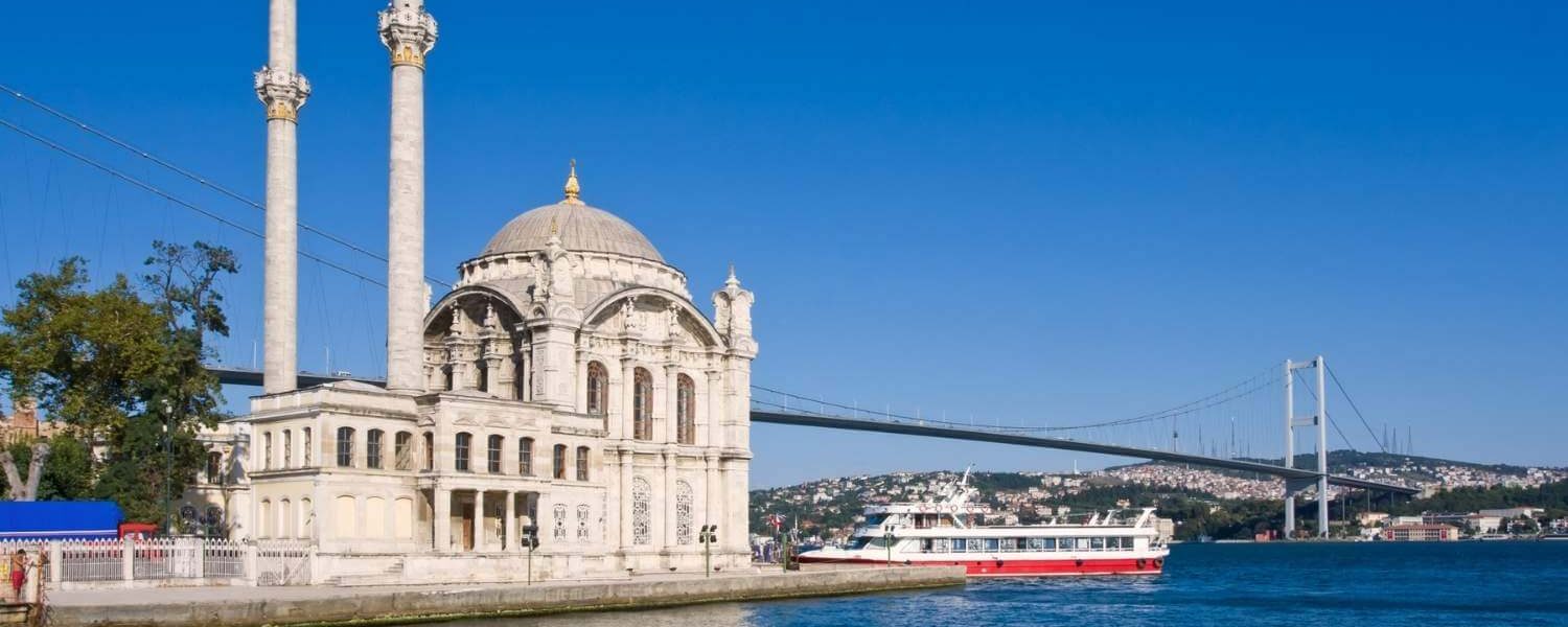 Ortaköy Mosque with twin minarets on the Bosphorus waterfront, with a ferry and the Bosphorus Bridge under a clear blue sky.