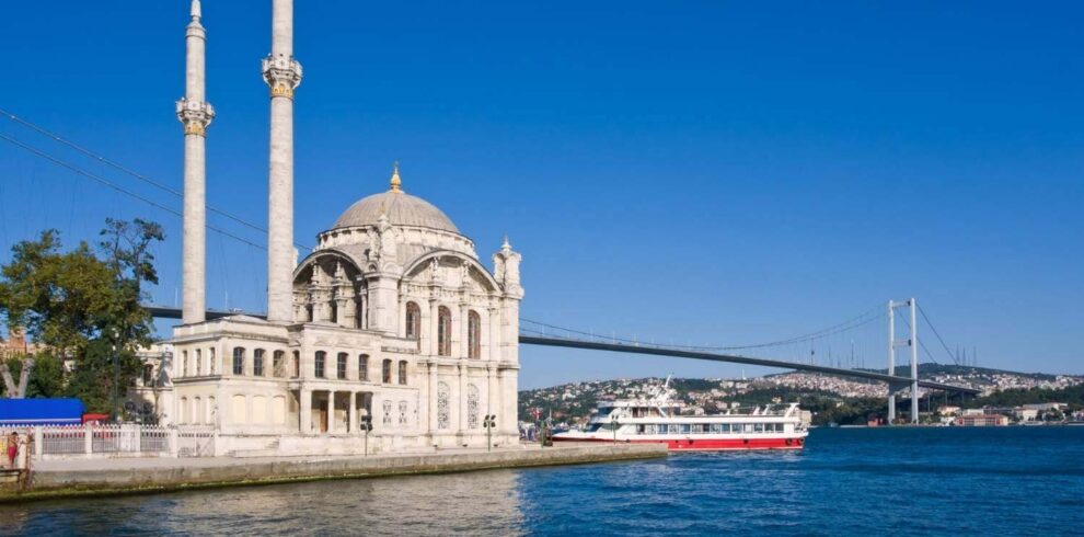 Ortaköy Mosque with twin minarets on the Bosphorus waterfront, with a ferry and the Bosphorus Bridge under a clear blue sky.