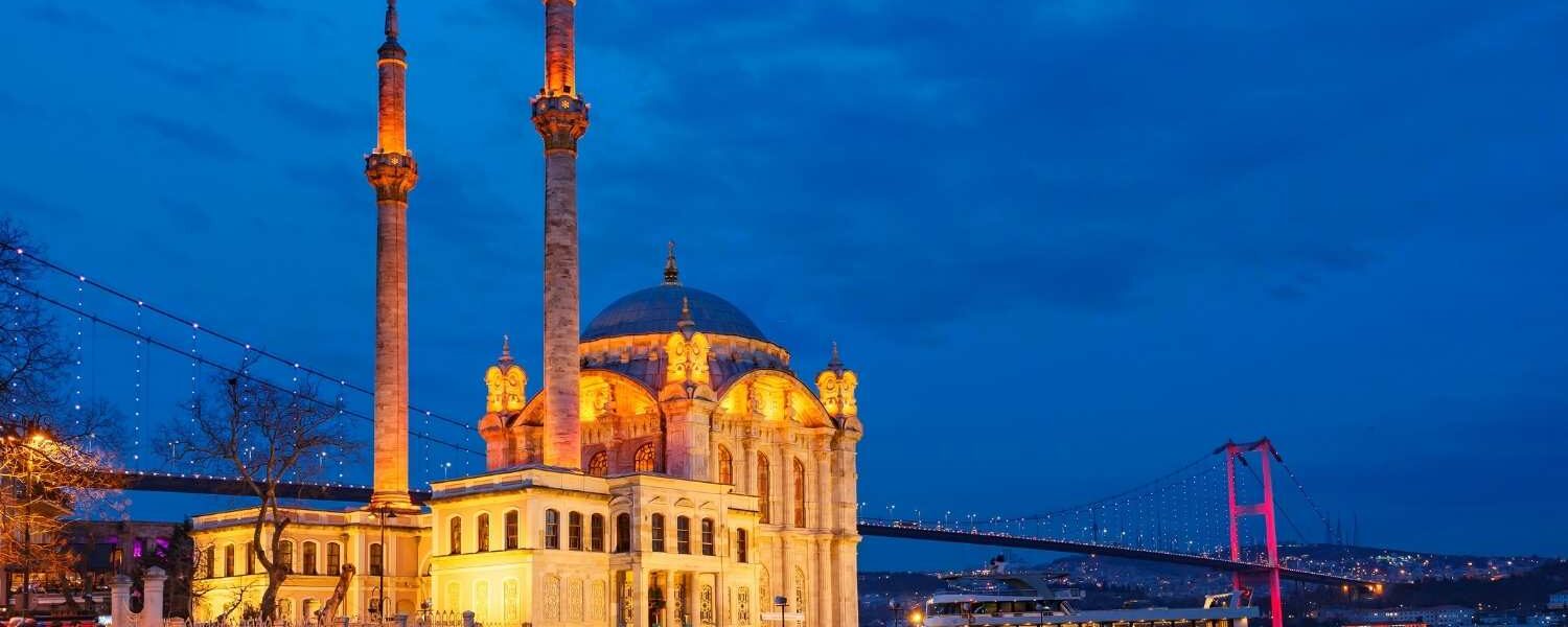 Ortaköy Mosque illuminated in warm lights at dusk, with the Bosphorus Bridge lit in red, reflecting on the water in Istanbul.