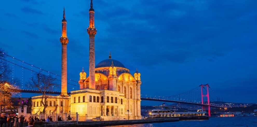 Ortaköy Mosque illuminated in warm lights at dusk, with the Bosphorus Bridge lit in red, reflecting on the water in Istanbul.