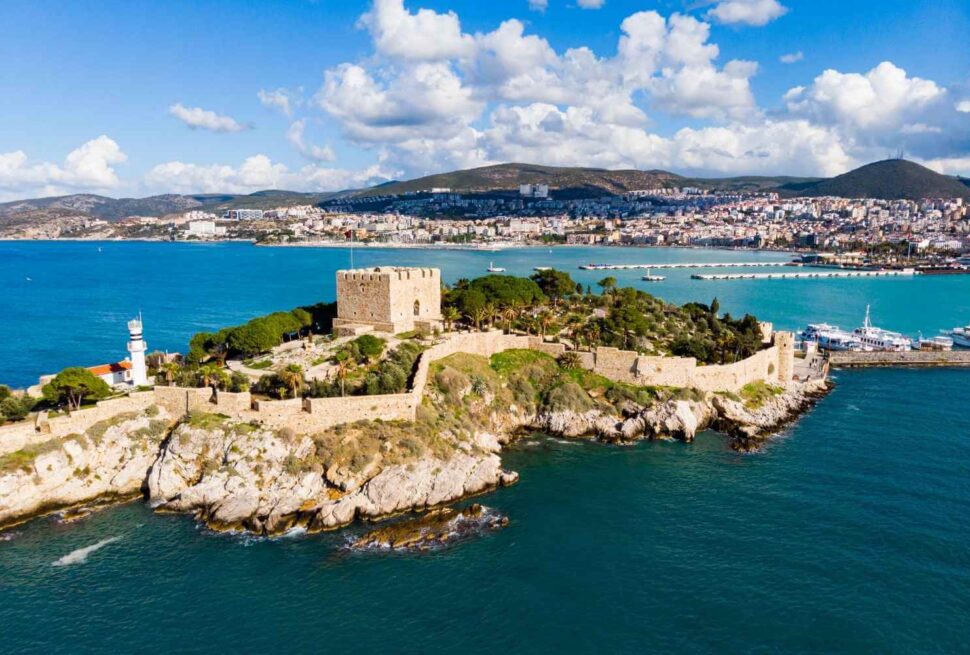 Aerial view of Kusadasi's scenic coastline featuring a historic castle on Pigeon Island surrounded by turquoise waters.